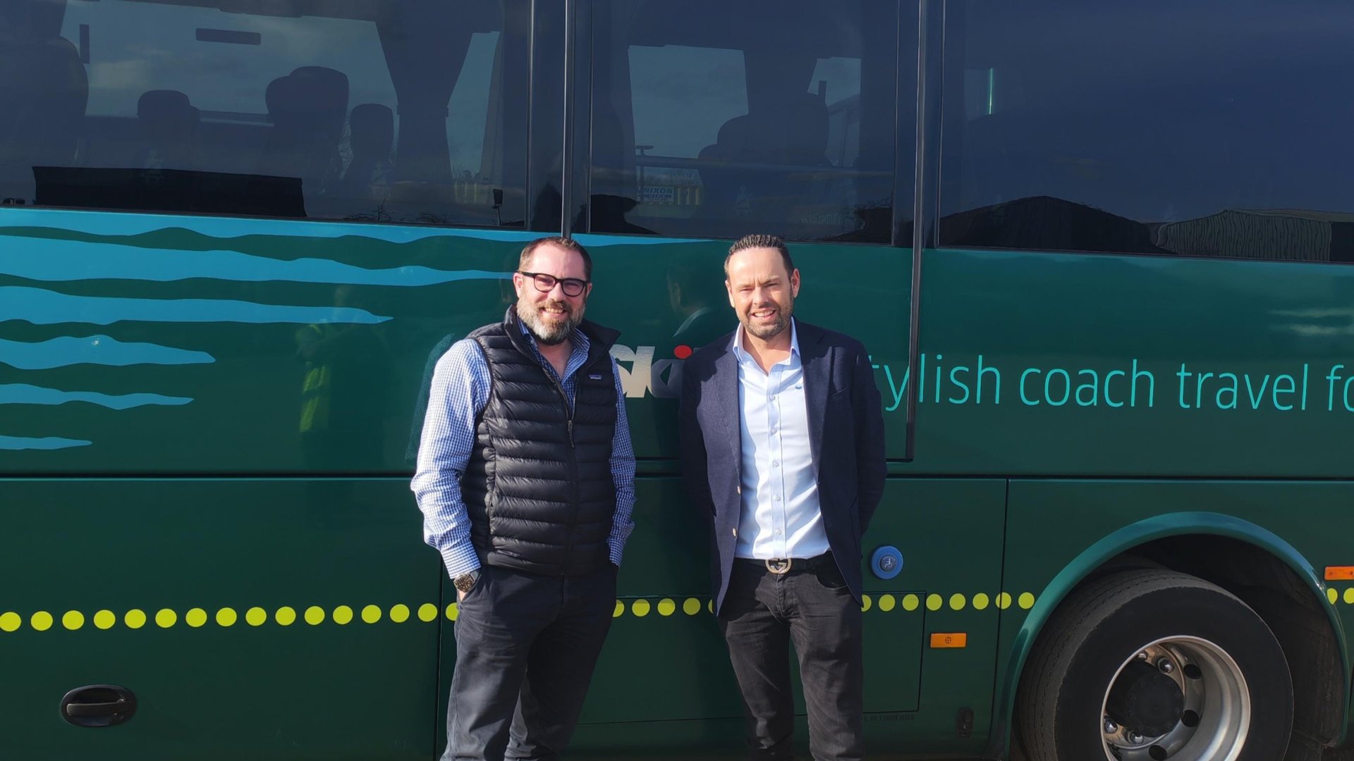 Two men standing beside a green coach with "stylish coach travel for" text, representing IT support for coach operators.
